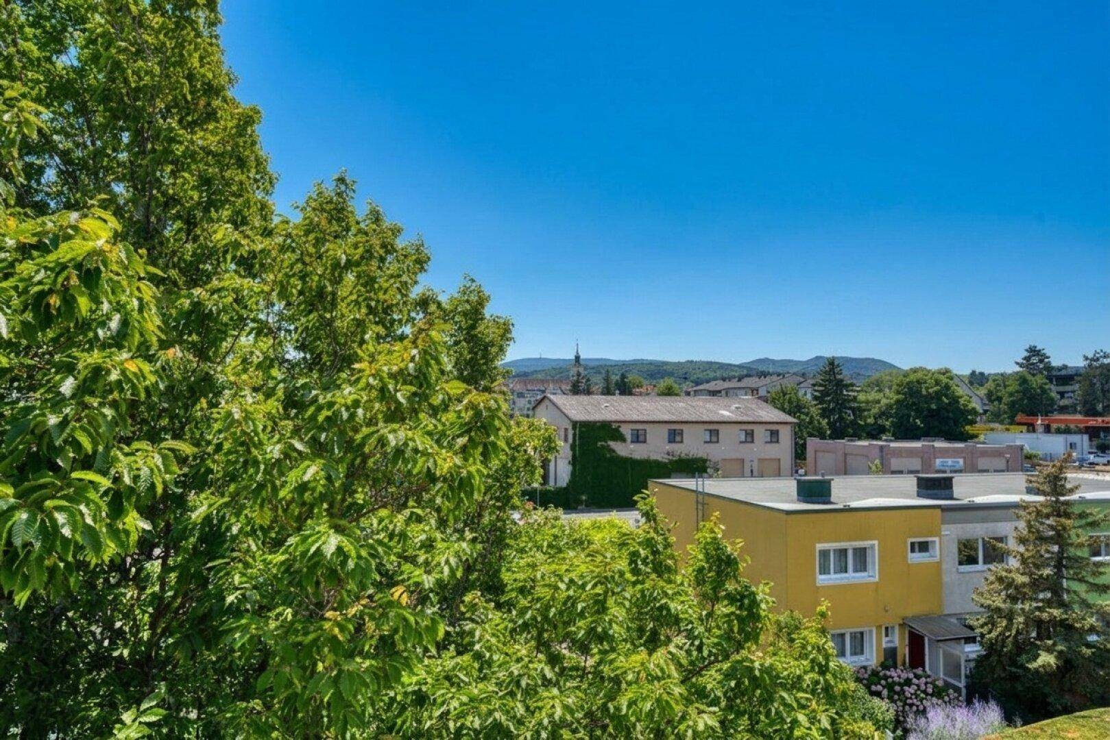 Weitblick über grüne Bäume auf Gebäude und ferne Hügel unter blauem Himmel.