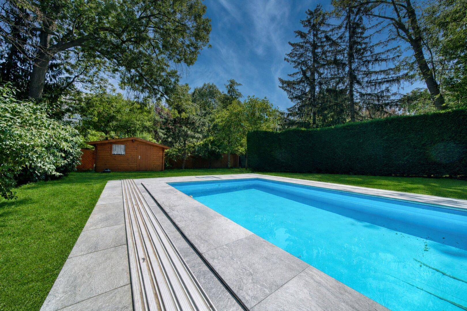 Langer Swimmingpool im Garten mit automatischer Abdeckung und Gartenhaus im Hintergrund.
