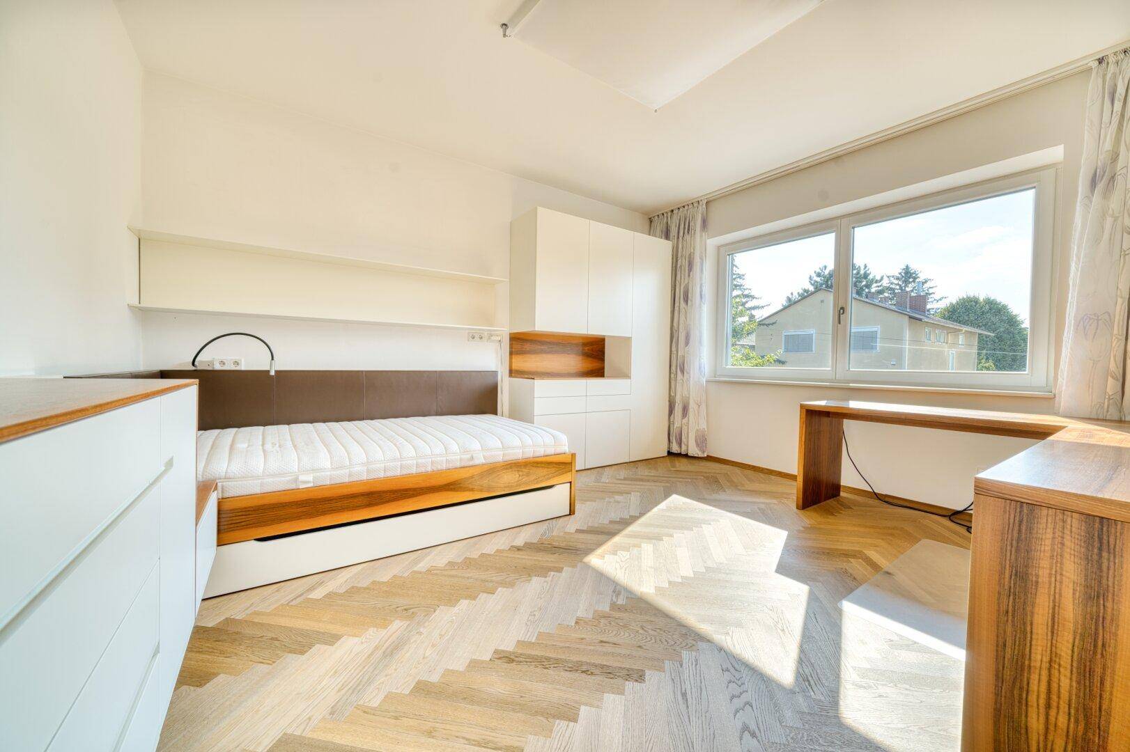 Helles Schlafzimmer mit Fischgrätparkett, Bett und Schreibtisch am Fenster.