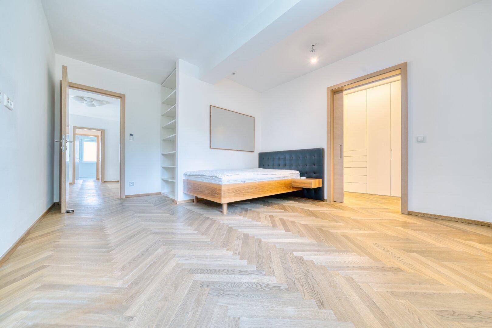Schlafzimmer mit Fischgrätparkett, Bettrahmen und integriertem Regal.