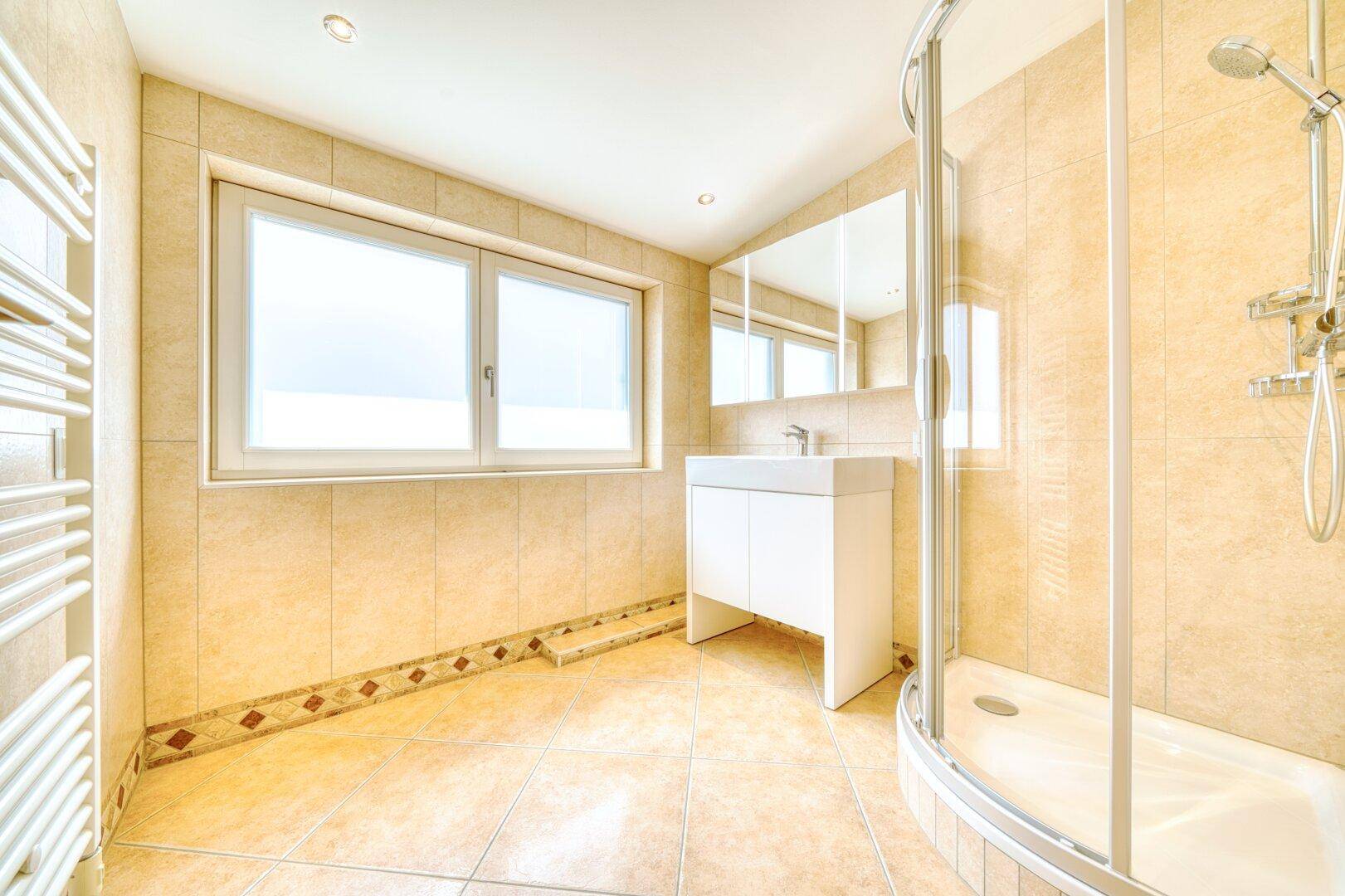 Modernes Badezimmer mit beigen Fliesen, Duschkabine, Waschbecken und Fenster.