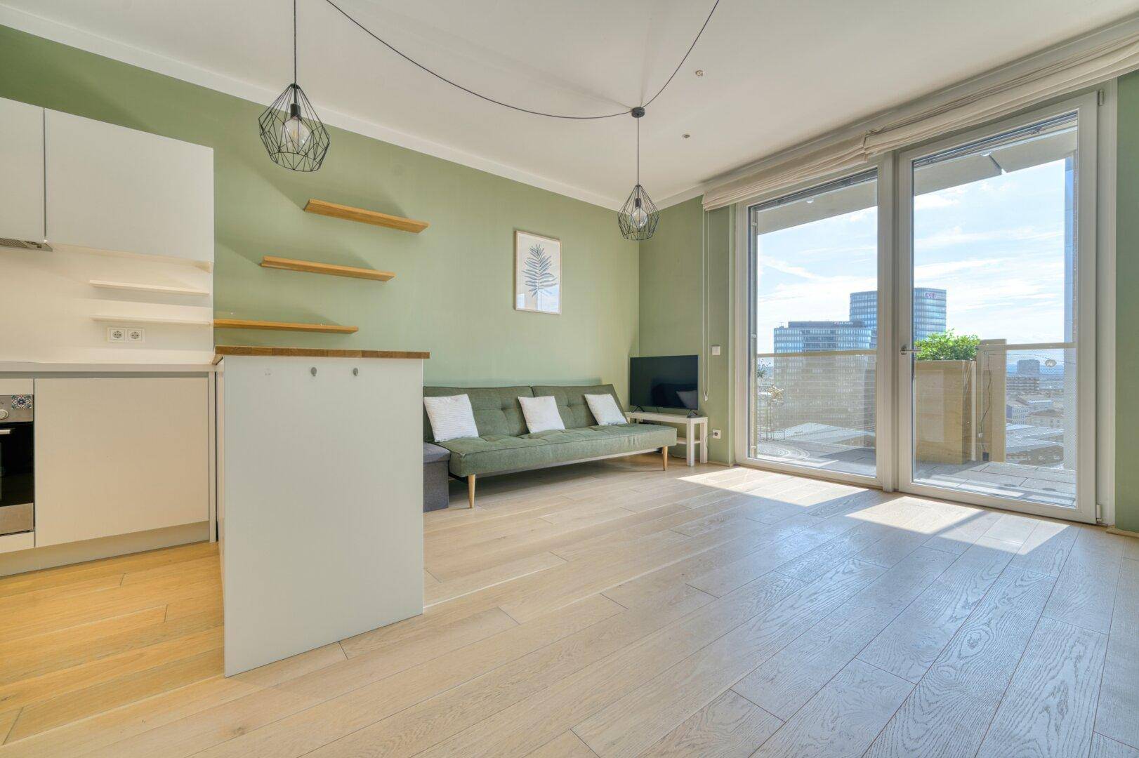 Heller offener Wohnbereich mit integrierter Küche, grün gestrichener Wand und Zugang zum Balkon.