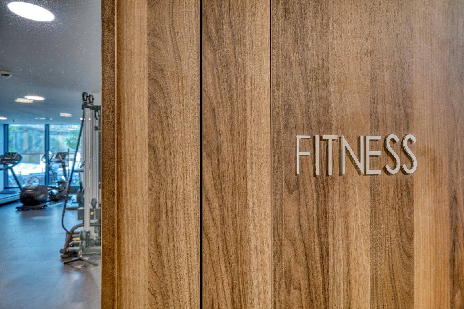 Holztür mit "FITNESS"-Schild, die den Eingang zu einem modernen Fitnessraum markiert.
