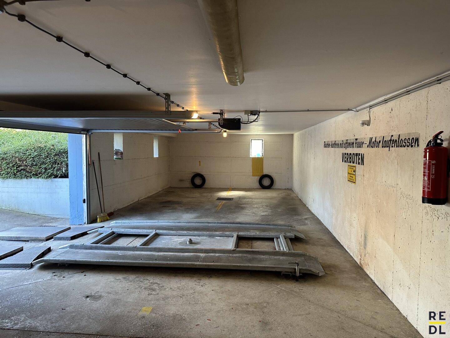 Innenansicht der Tiefgarage mit Autoaufzug, Feuerlöscher und Hinweisschildern an der Wand.