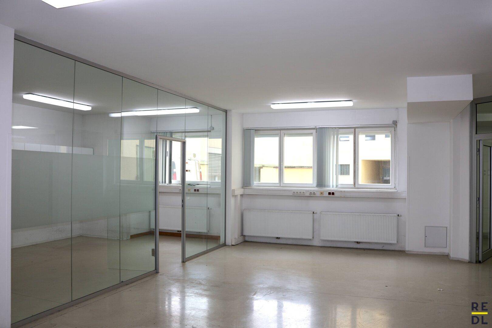 Helles, leeres Büro mit Glaswänden und großen Fenstern, die viel Tageslicht hereinlassen.