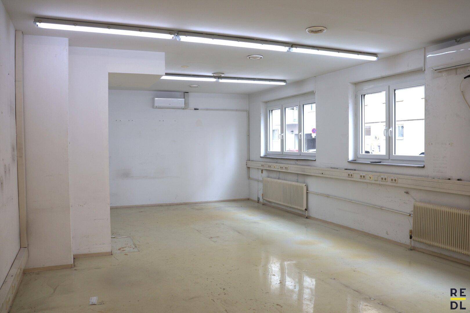 Leerer Büroraum mit Fenstern, Heizkörpern und sichtbaren Klimaanlagen an der Wand.