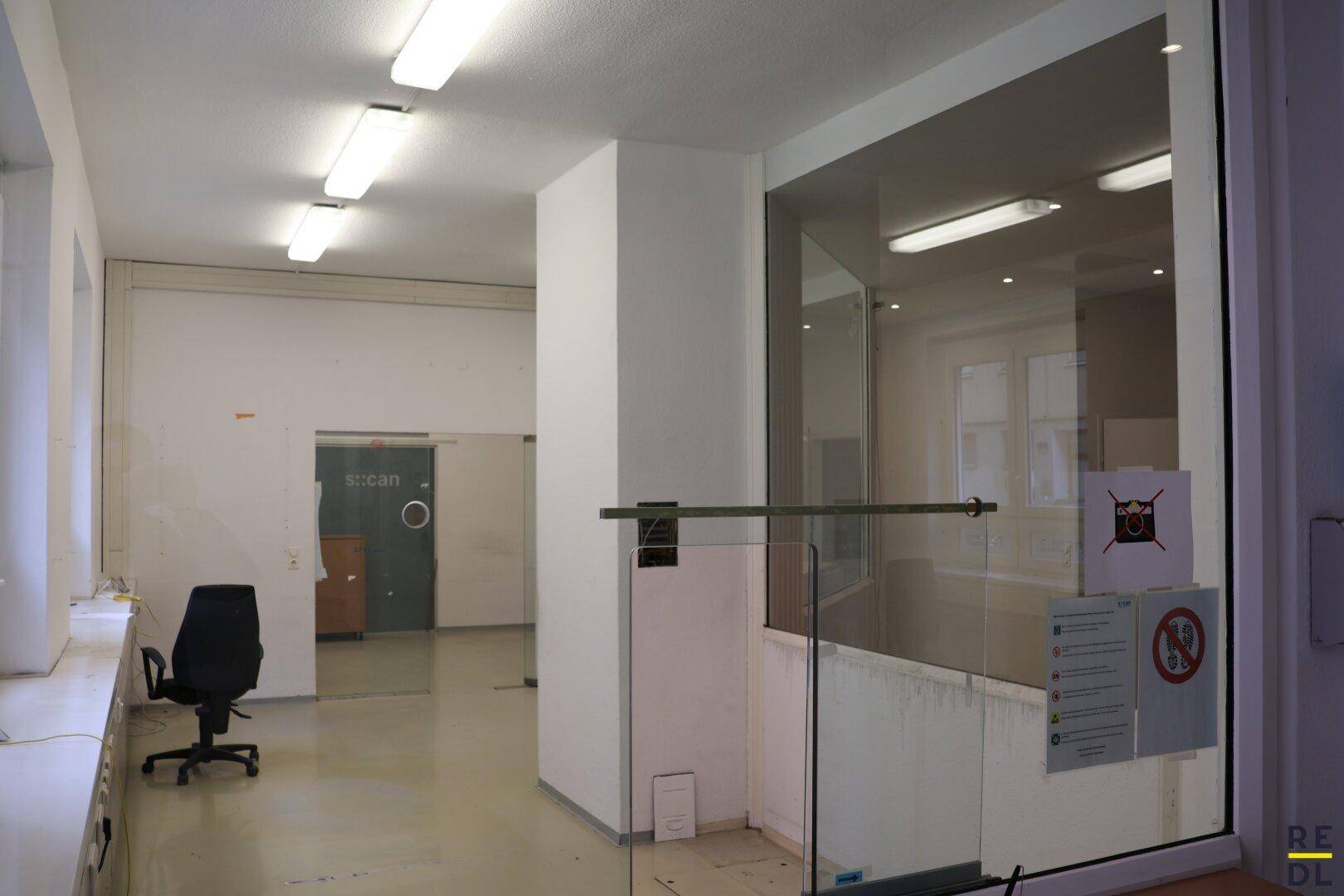 Bürobereich mit Glasabtrennung, einem Bürostuhl und Blick in einen weiteren Raum.