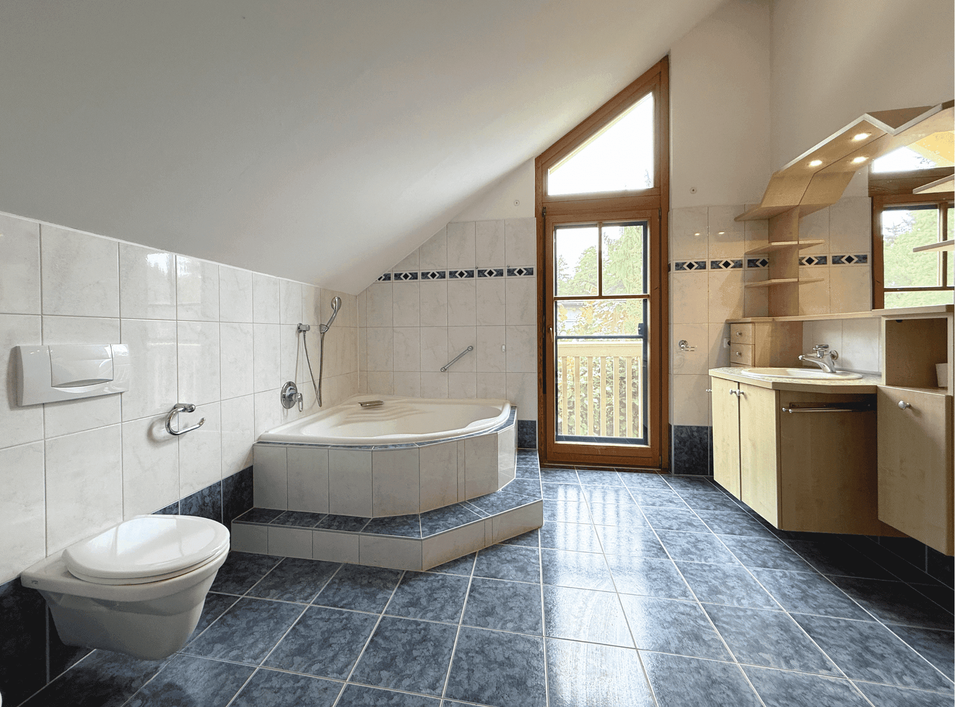 Badezimmer im Dachgeschoss mit blauen Fliesen, Eckbadewanne und Fenster mit Ausblick.