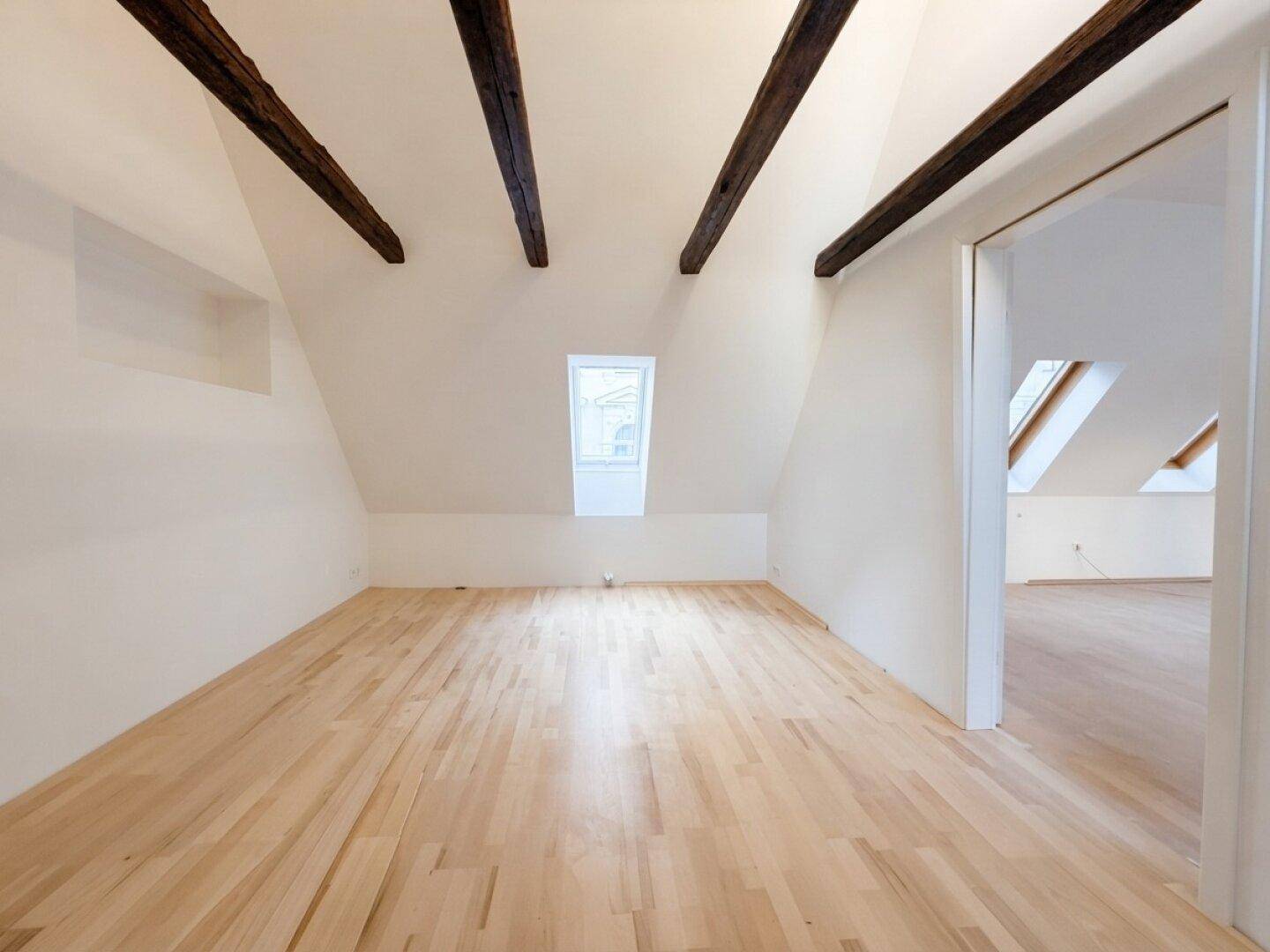 Dachgeschossraum mit sichtbaren Holzbalken, hellem Holzboden und einem Dachfenster.