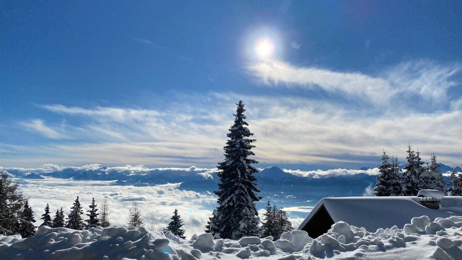 Winterliche Berglandschaft mit tief verschneiten Bäumen und einem sonnigen Himmel über den Wolken.