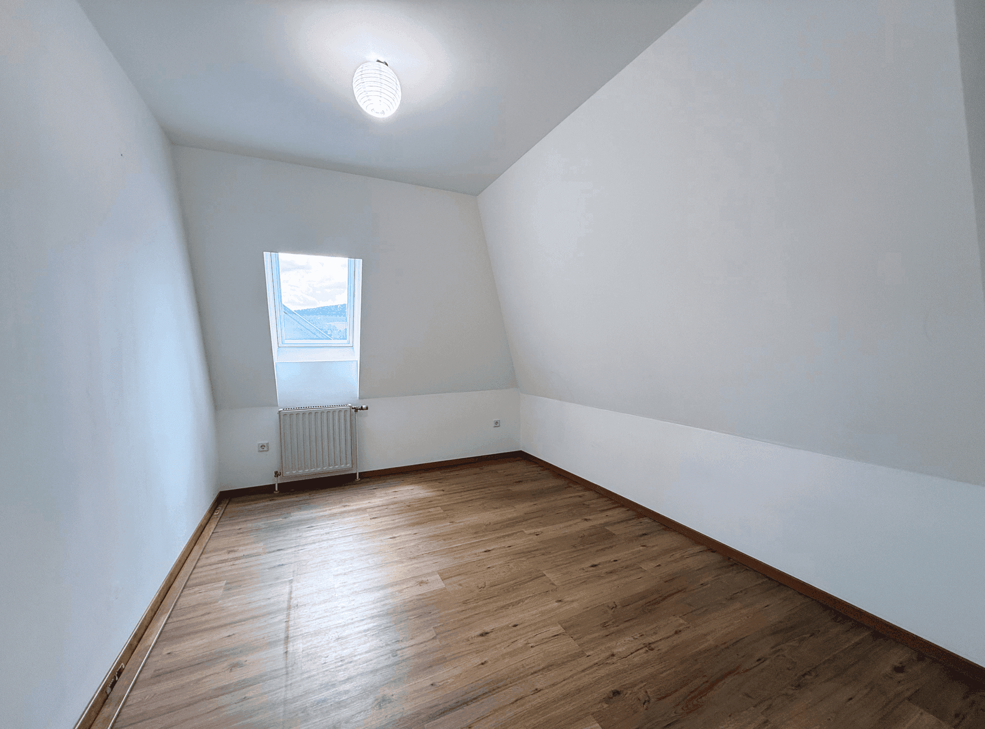 Freundliches Dachzimmer mit Laminatboden, weiß gestrichenen Wänden und einem großen Dachfenster.
