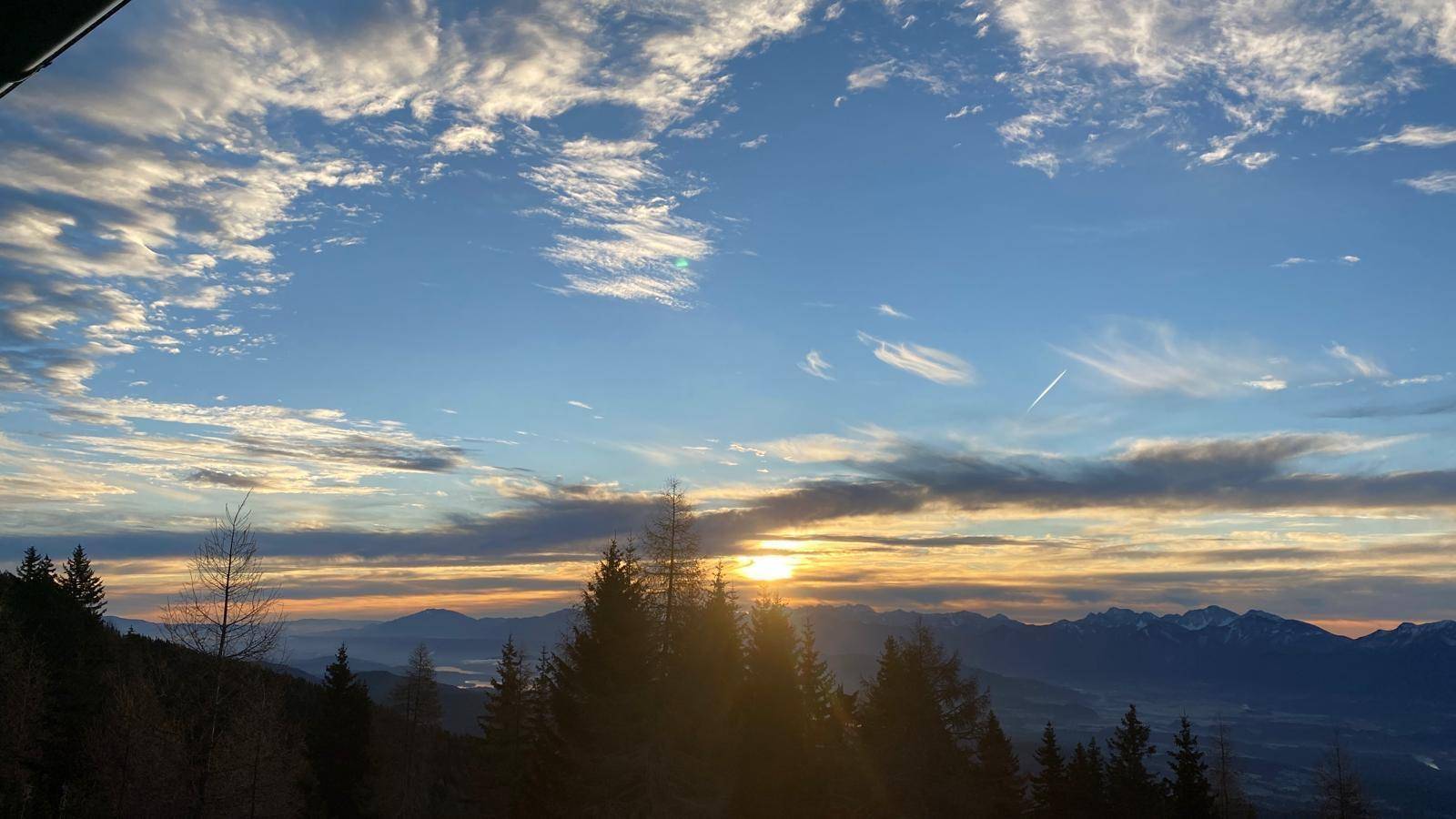 Atemberaubender Sonnenuntergang über einer Bergkette mit silhouettenhaften Bäumen im Vordergrund.