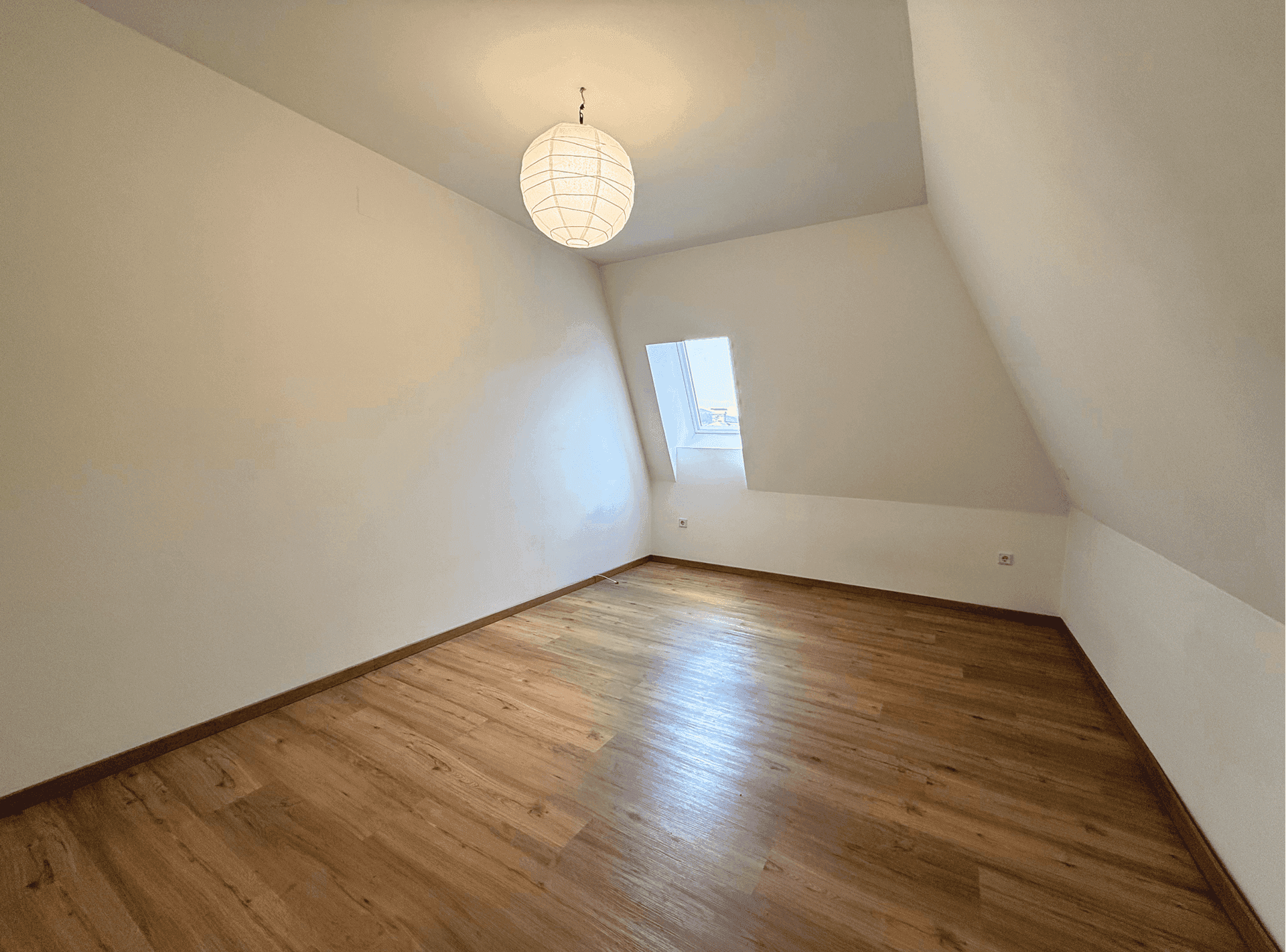 Helles Dachzimmer mit schrägen Wänden, Laminatboden und einem Dachfenster für natürliches Licht.