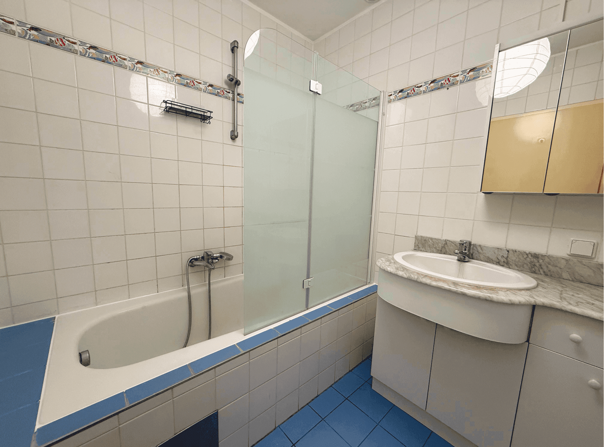 Badezimmer mit Badewanne, Duschabtrennung, Waschtisch und blauen Bodenfliesen im 80er-Jahre-Stil.