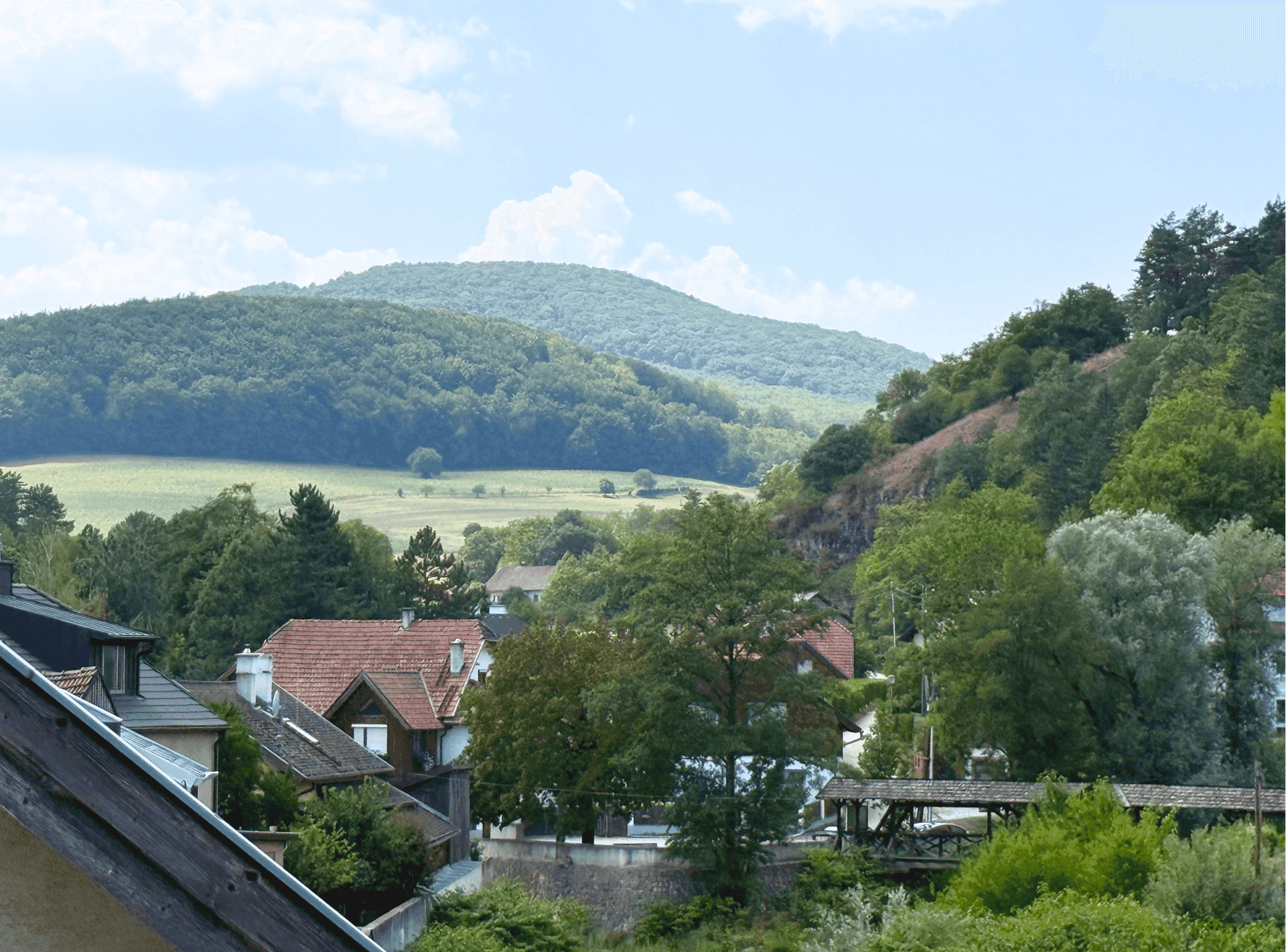 Weitläufiger Blick über eine grüne Hügellandschaft mit Häusern und Bäumen unter blauem Himmel.