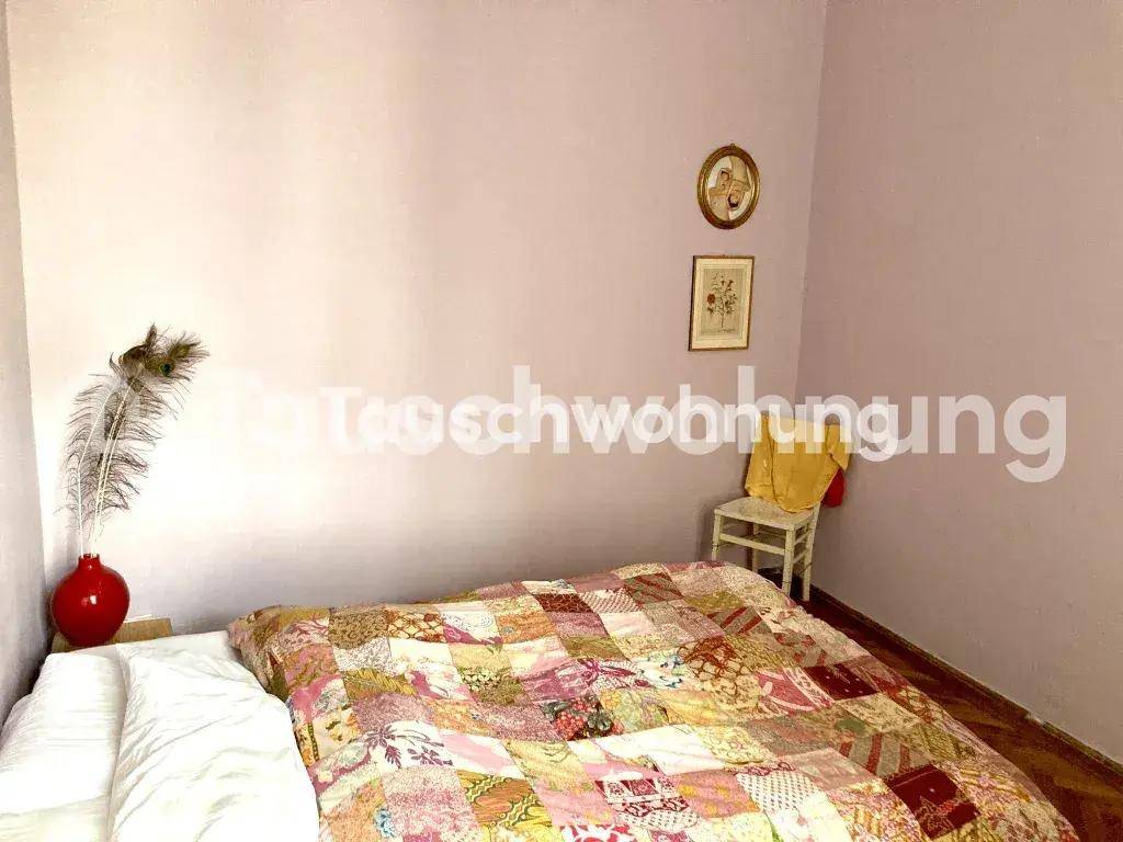 Schlafzimmer mit Bett, bunter Tagesdecke, hellvioletten Wänden und einem kleinen Stuhl.