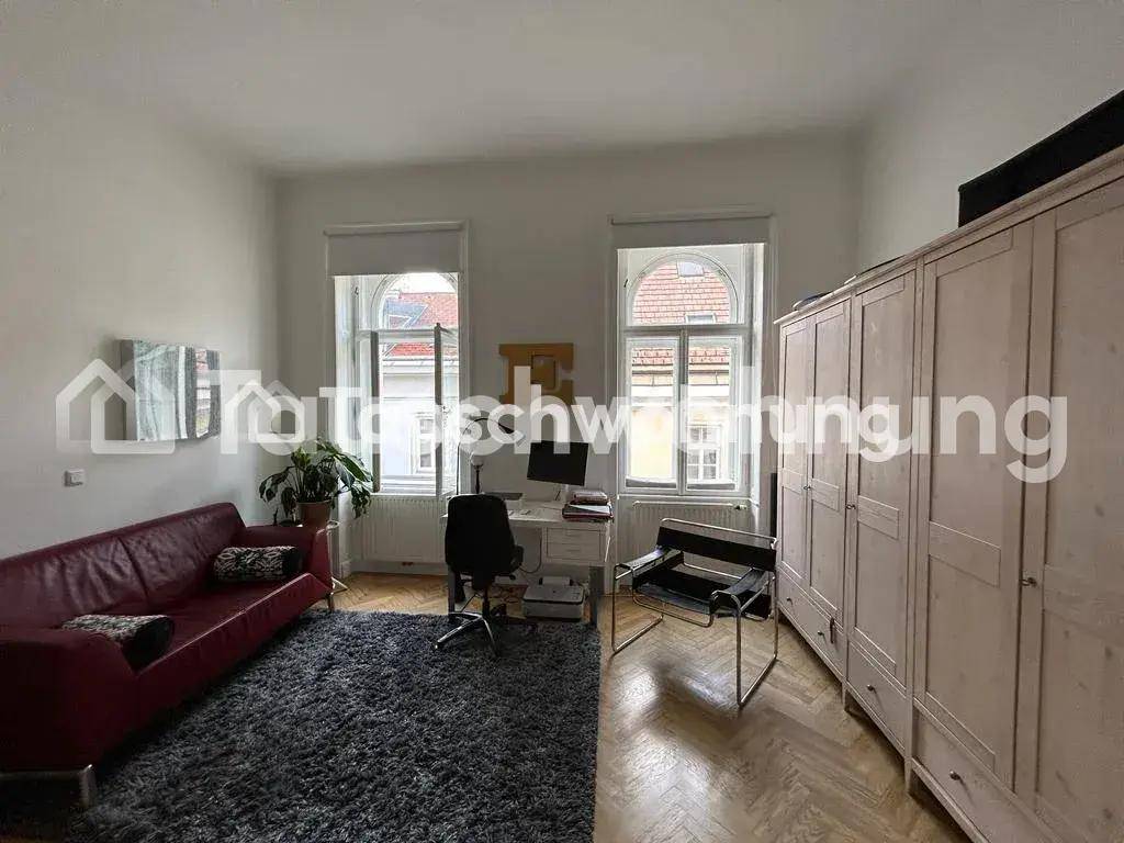 Wohnzimmer mit rotem Sofa, Schreibtisch und großem Kleiderschrank, beleuchtet durch hohe Fenster.