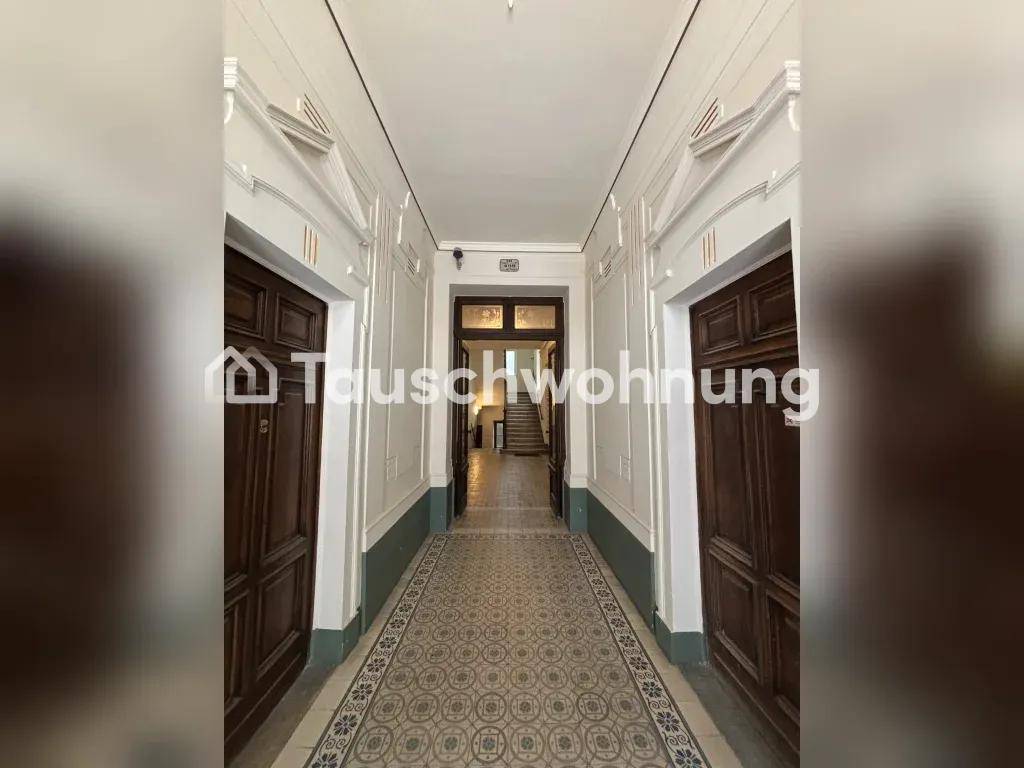 Langer Flur in einem Altbau mit gemusterten Fliesen, hohen Decken und Holztüren, der zum Treppenhaus führt.