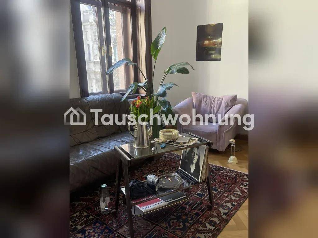 Helles Wohnzimmer mit Ledersofa, bequemem Sessel, Glastisch und großer Zimmerpflanze auf gemustertem Teppich.