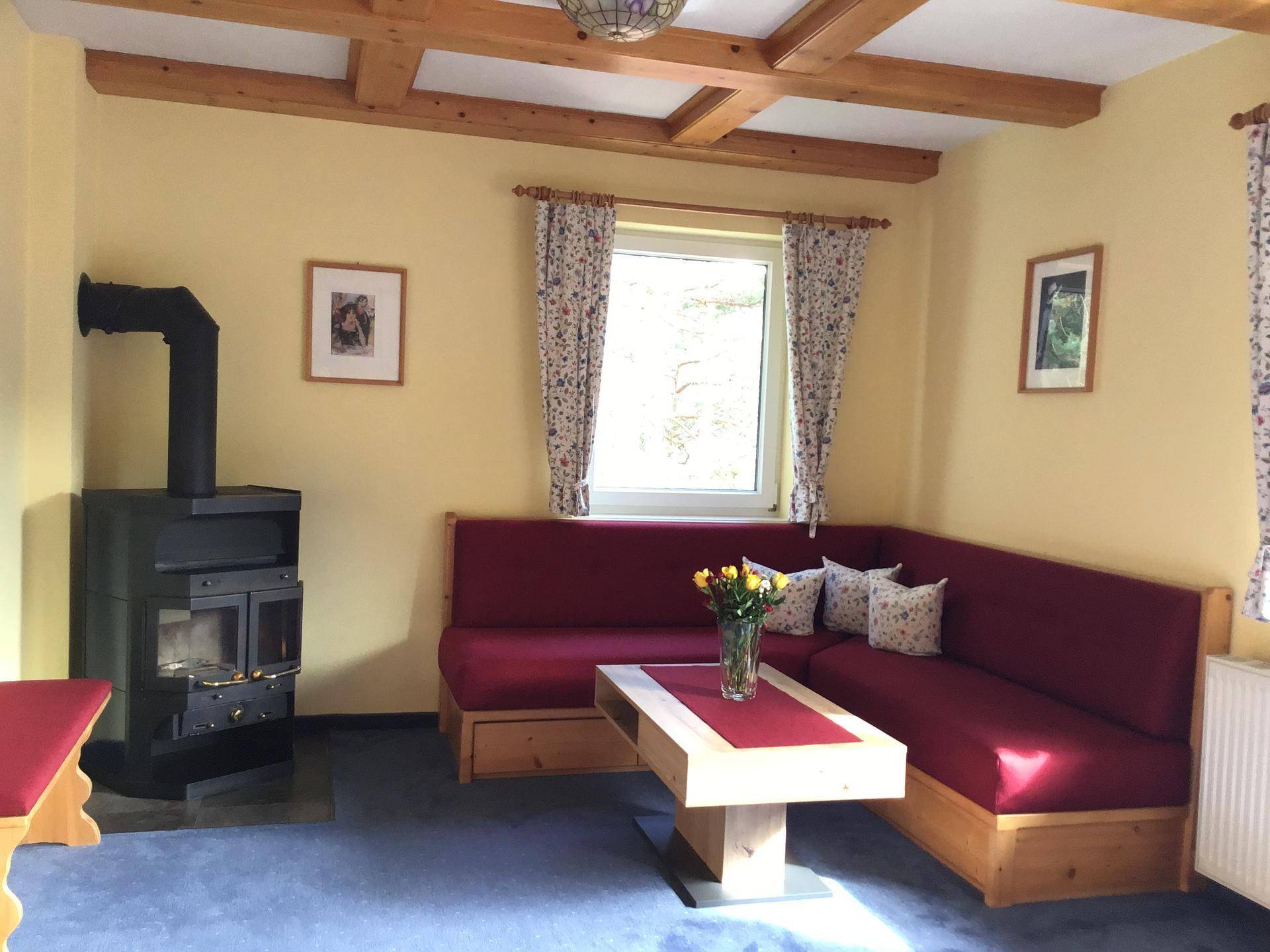 Rustikales Wohnzimmer mit Kaminofen, gemütlicher Sitzecke und sichtbaren Holzdeckenbalken.