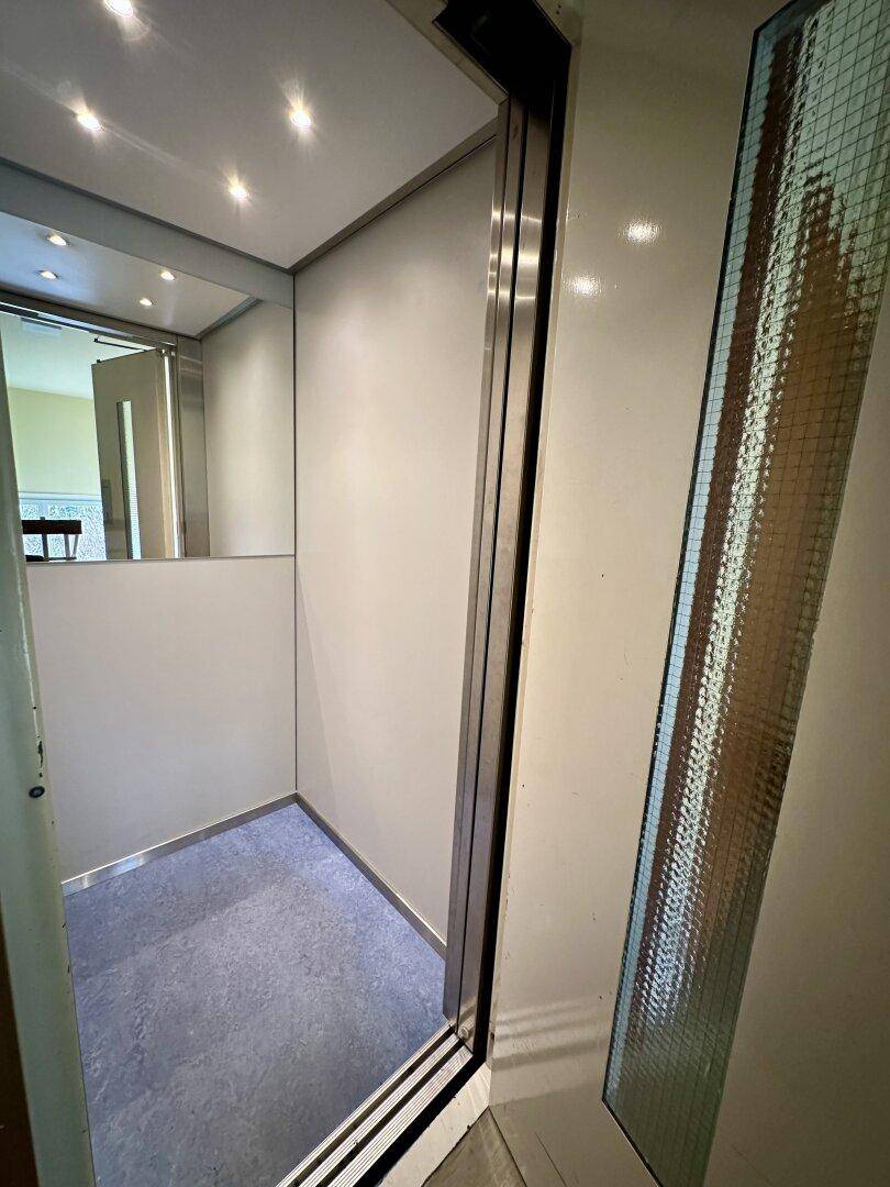 Moderner Aufzug mit Spiegelfläche und blauer Bodenmatte, zugänglich für alle Etagen.