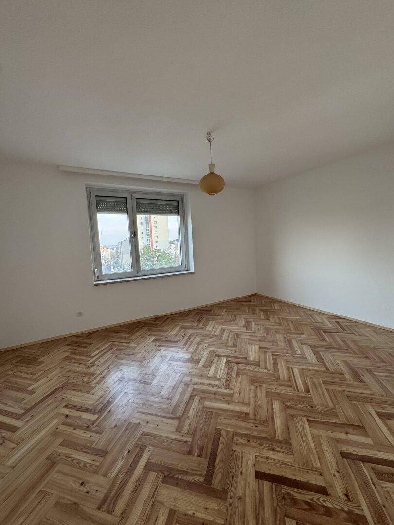 Geräumiger Raum mit Fischgrätparkett, weißen Wänden und einem Fenster mit Rollladen.