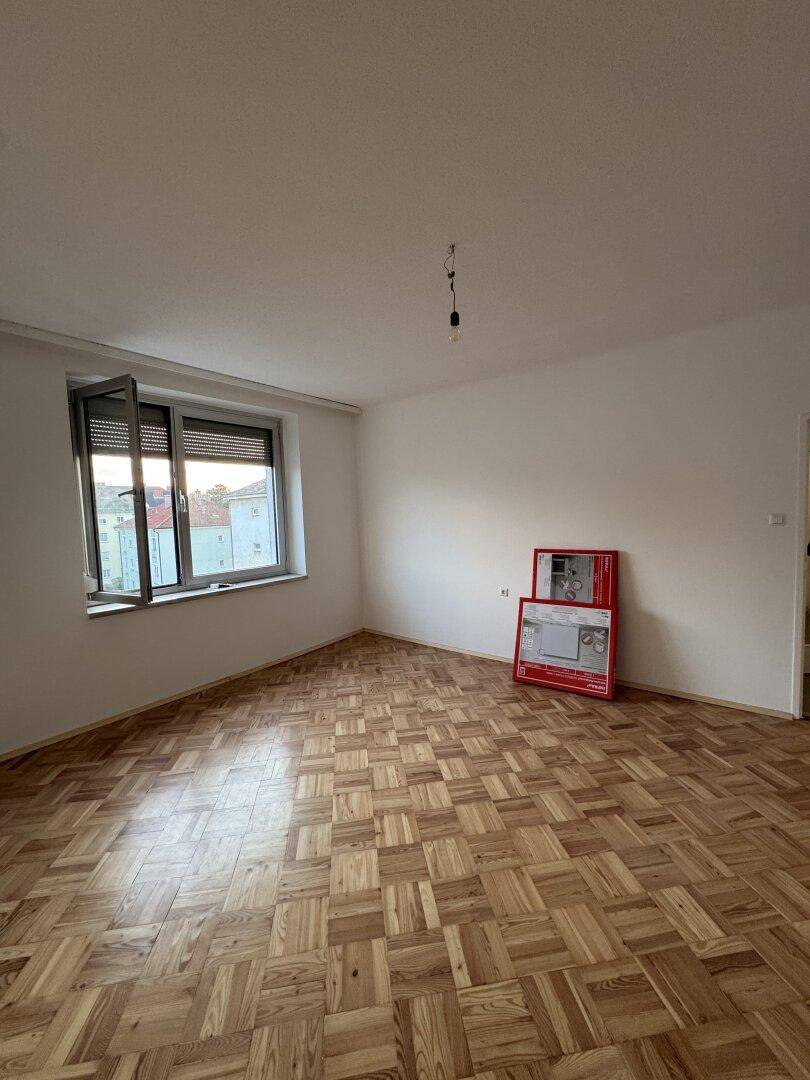 Leerer Raum mit Parkettboden, weißen Wänden und einem Fenster, das viel Licht hereinlässt.