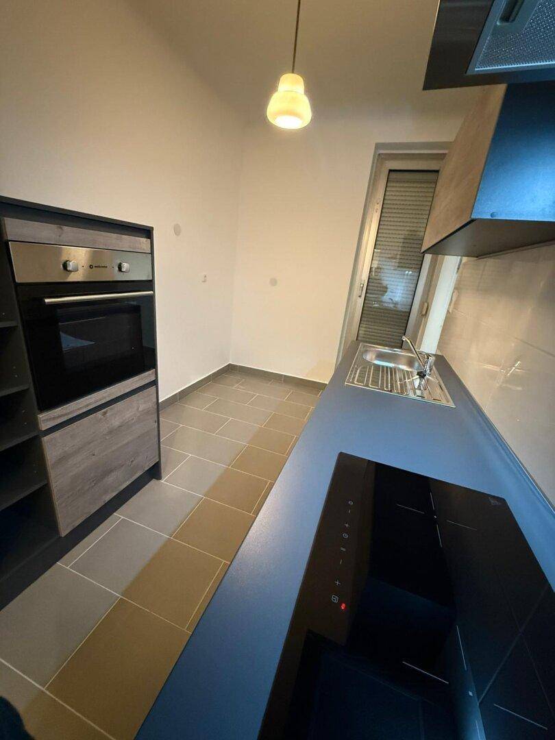 Blick in die Küche mit Backofen, Induktionskochfeld und Zugang zu einem Fenster mit Rollladen.