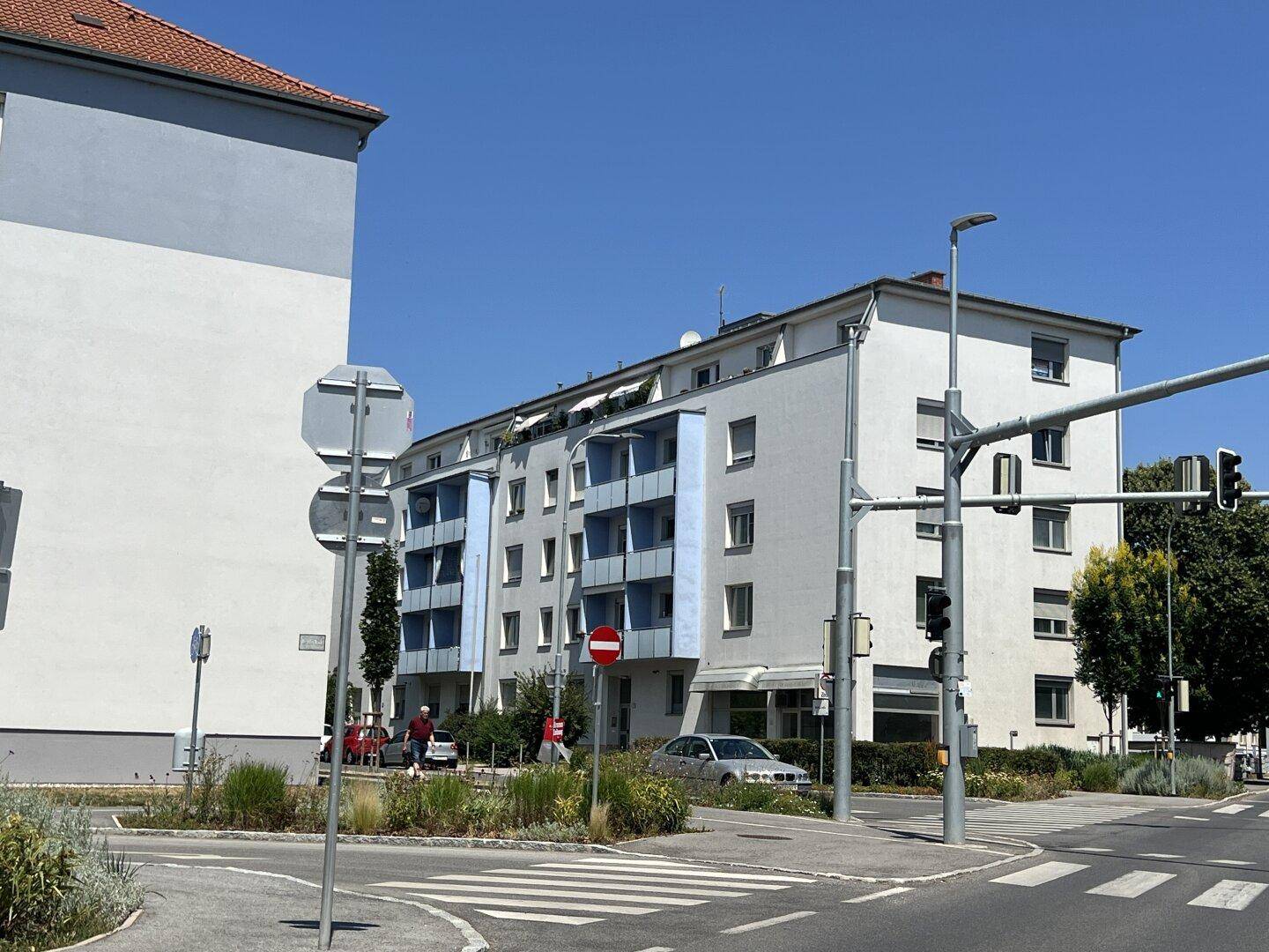 Mehrfamilienhaus mit blauen Balkonen an einer sonnigen Straßenecke.