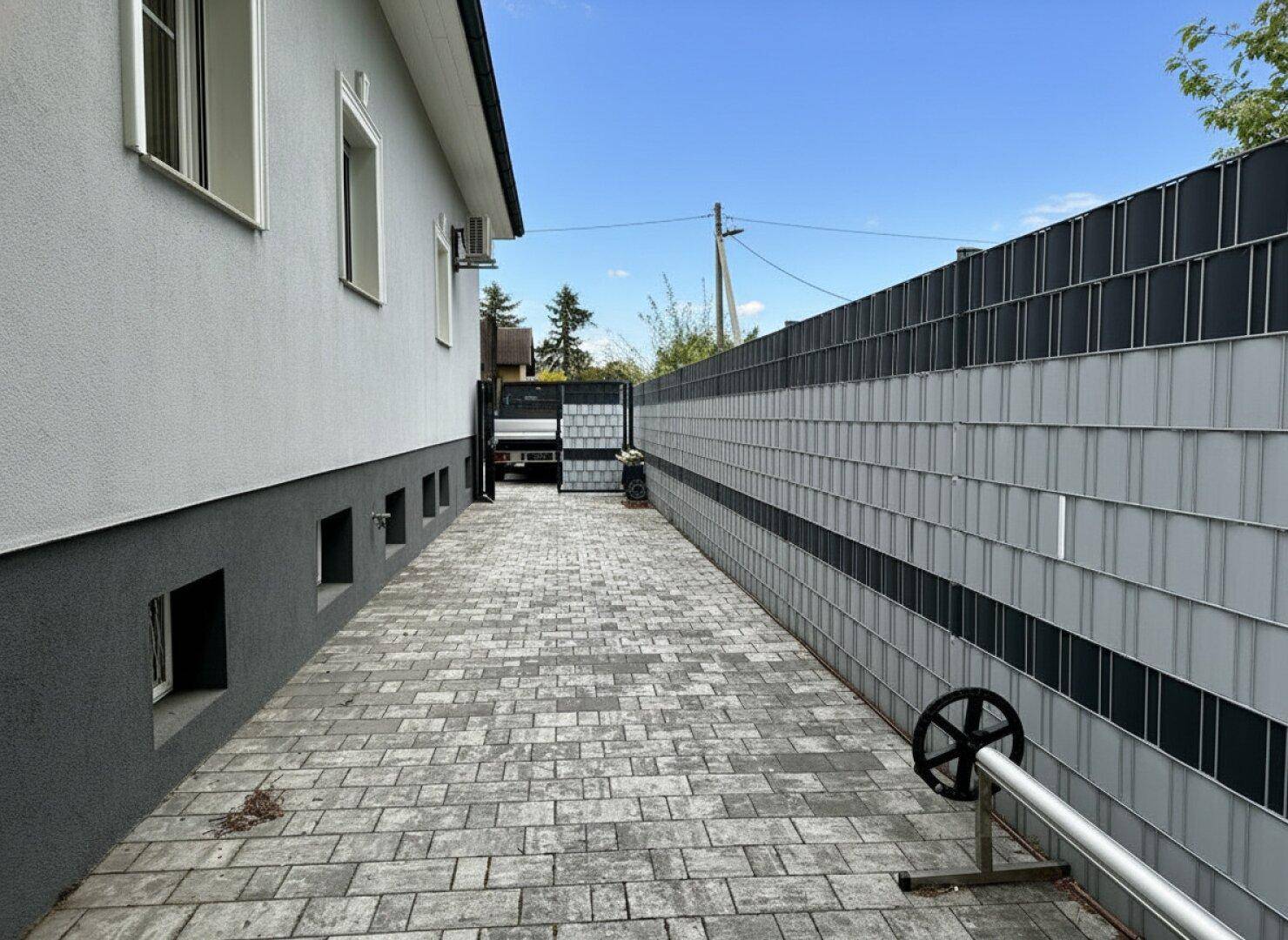 Gepflasterter Seitenweg entlang des Hauses mit Zaun und Zugang zum hinteren Bereich.