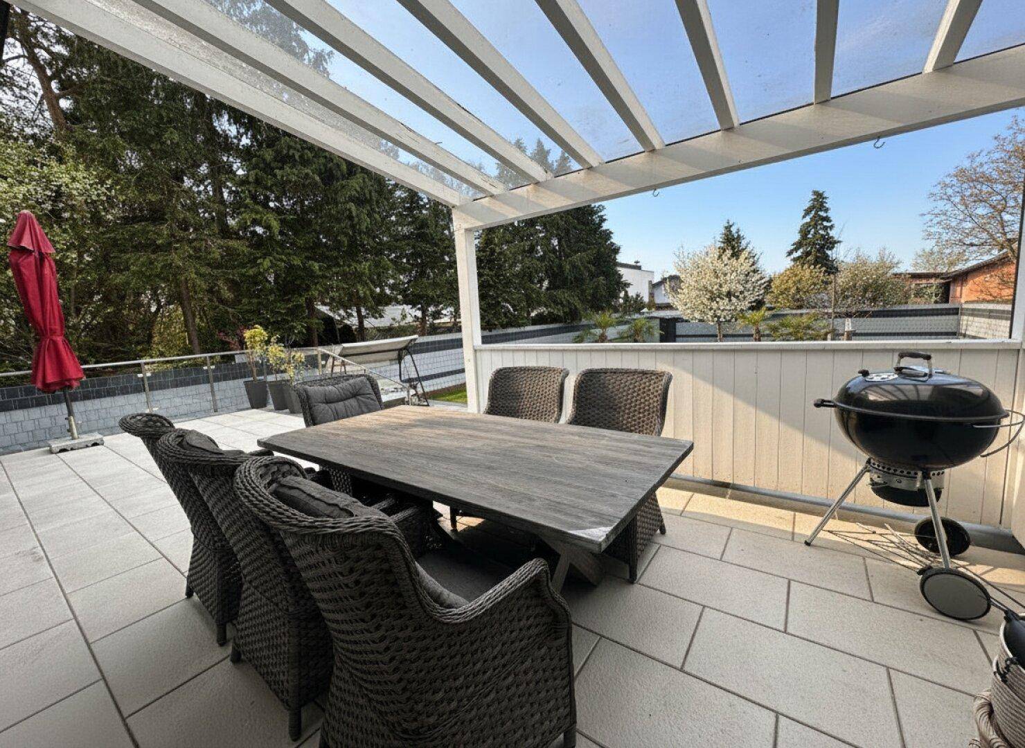Große überdachte Terrasse mit Rattanmöbeln und Grill, umgeben von Bäumen.
