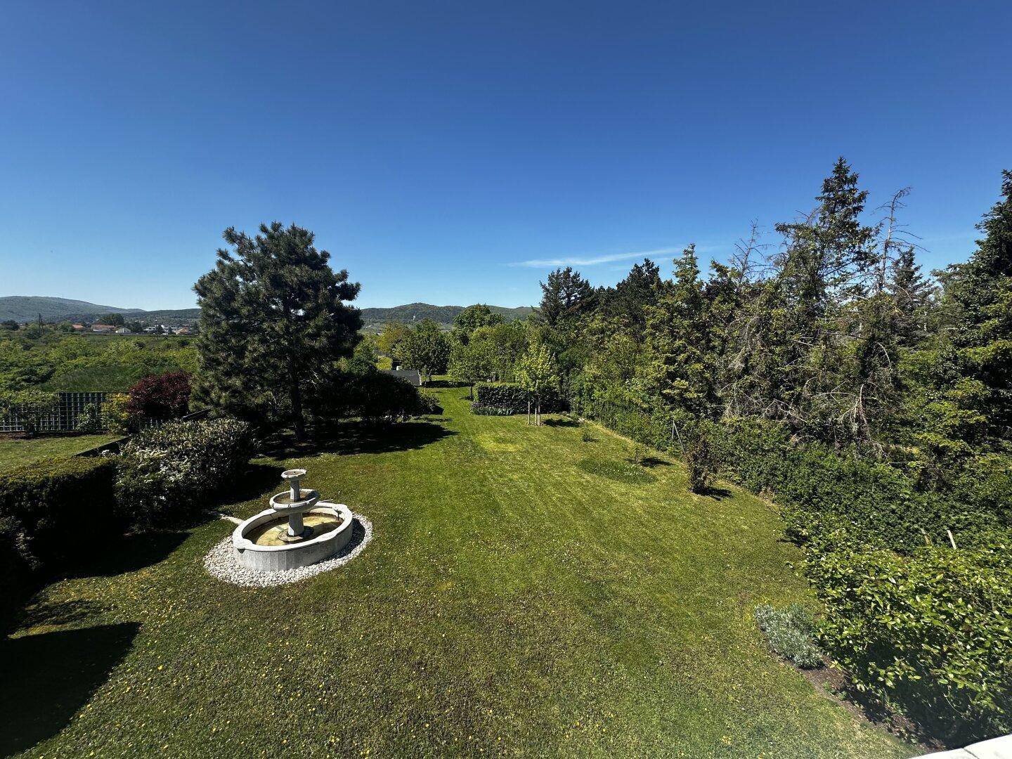 Weitläufiger Garten mit Rasenfläche, dekorativem Brunnen und altem Baumbestand.