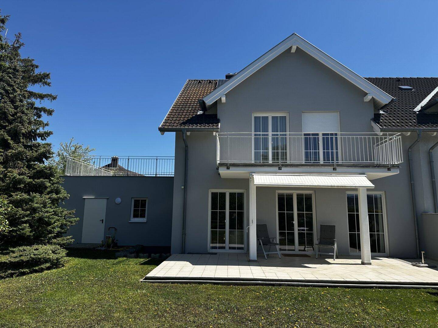 Rückansicht des Hauses mit grauer Fassade, großer Terrasse und Balkon im Obergeschoss.