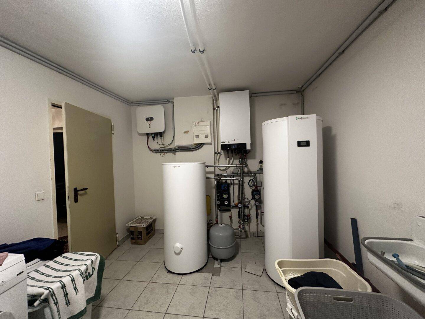 Technikraum mit zwei großen Warmwasserspeichern, Waschmaschine und Waschbecken auf Fliesenboden.