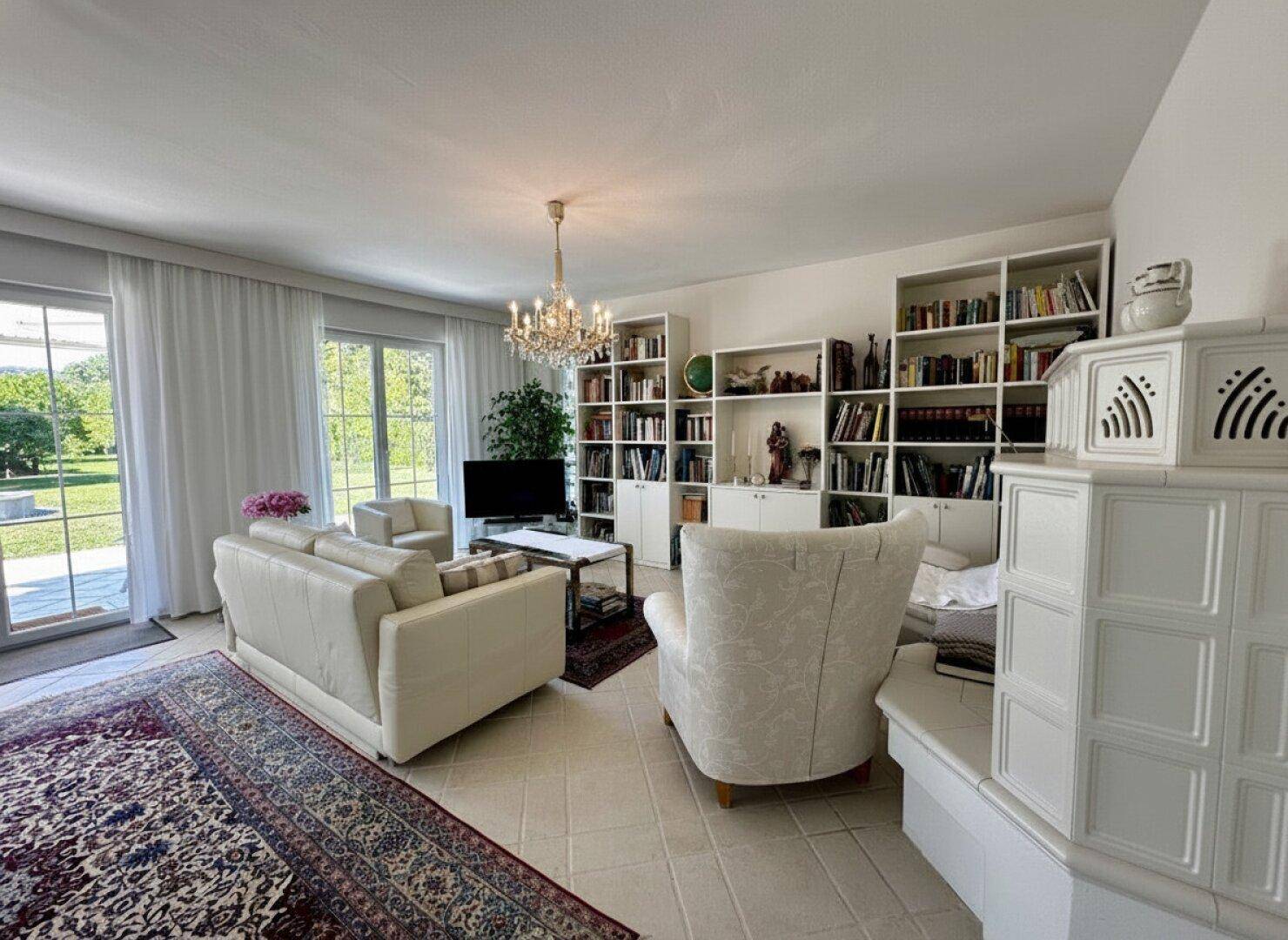 Gemütliches Wohnzimmer mit Kachelofen, Bücherregalen und Blick in den Garten.