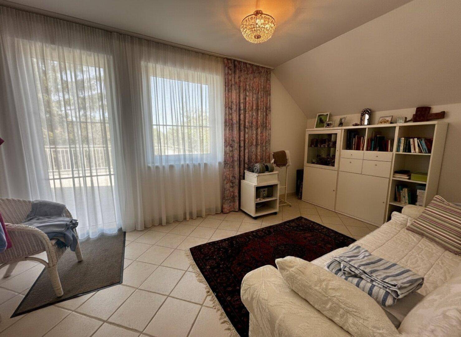 Helles Zimmer mit Fliesenboden, Sofa und großen Fenstern mit Blick ins Grüne.