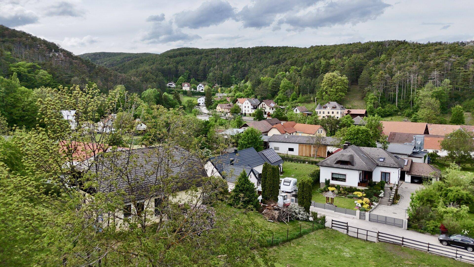 Luftaufnahme eines malerischen Dorfes in einem Tal, umgeben von bewaldeten Hügeln und vielen Wohnhäusern.