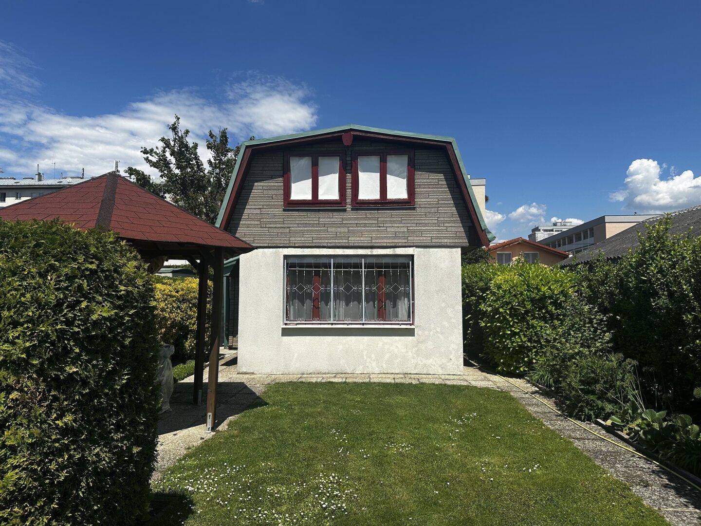 Kleines Haus mit weißer Fassade, Mansarddach und einem Pavillon im Vorgarten.