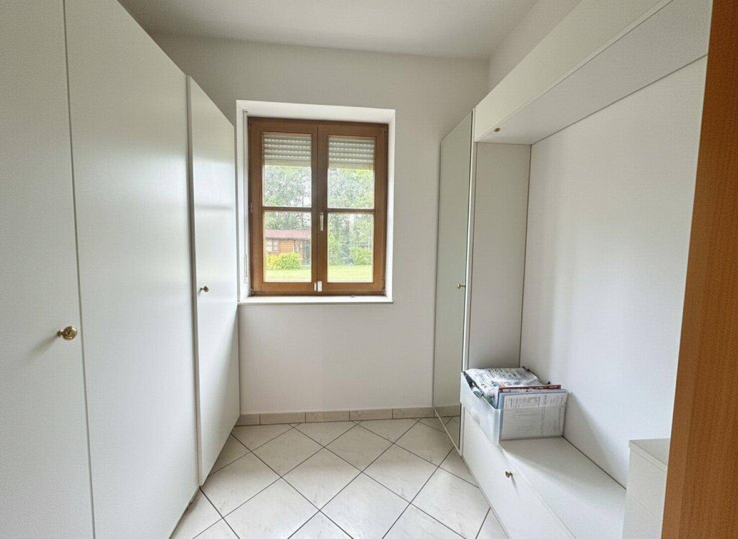 Heller Raum mit weißen Einbauschränken, Fliesenboden und Fenster mit Rollläden.
