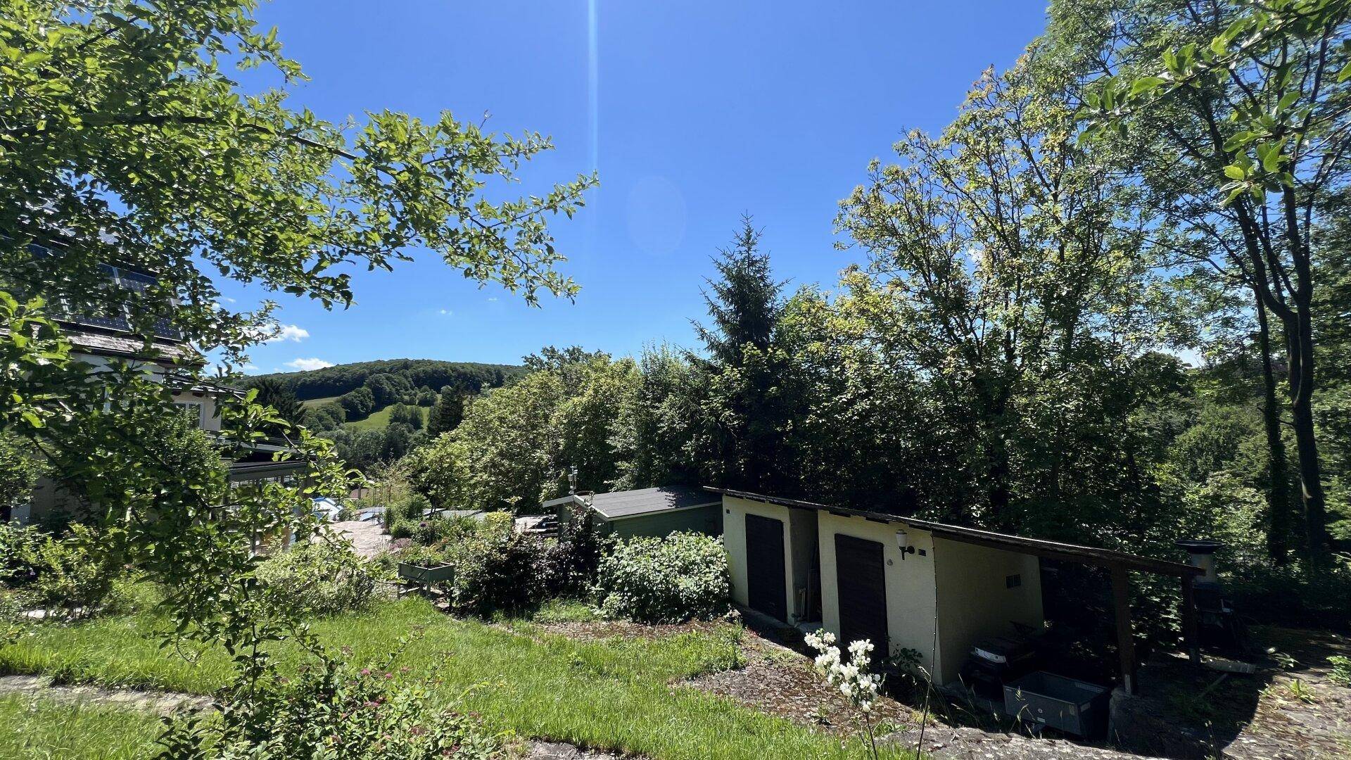 Gartenansicht mit einem kleinen Nebengebäude und weitem Blick auf bewaldete Hügel in der Ferne.