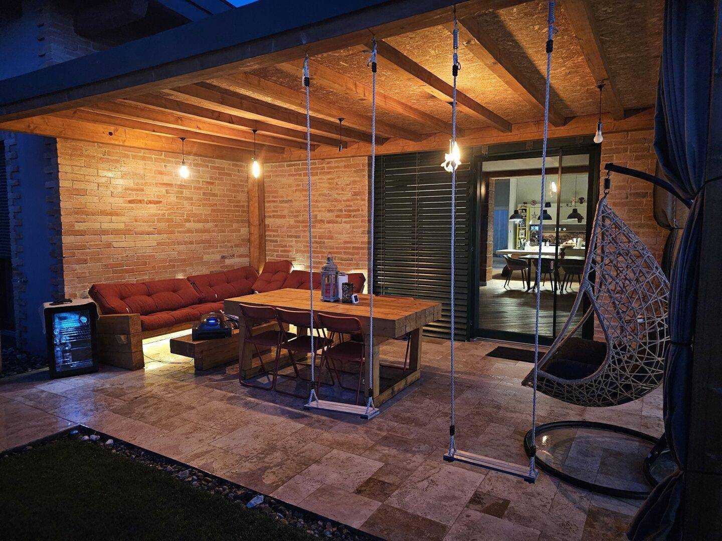 Beleuchtete überdachte Terrasse mit Esstisch, Schaukeln und Blick ins Haus bei Nacht.