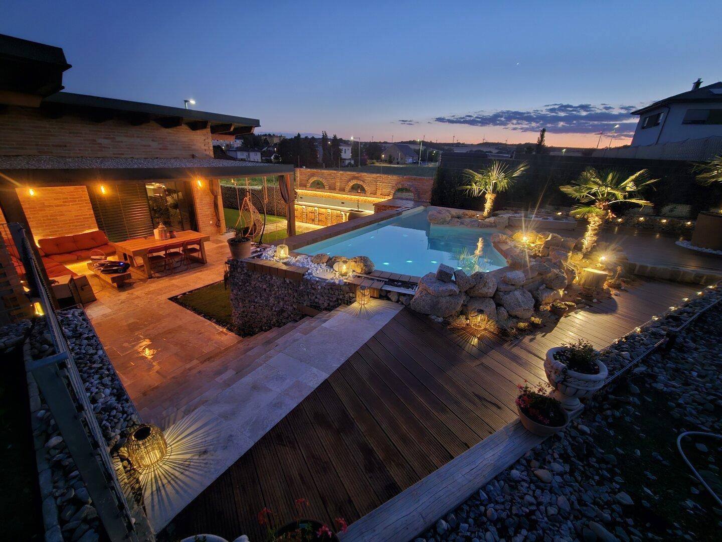 Panoramablick auf das beleuchtete Haus, den Pool und die Außenbereiche in der Abenddämmerung.