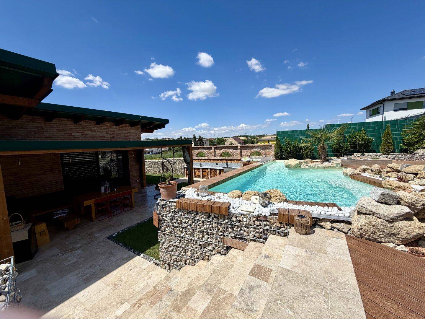 Weitläufiger Außenbereich mit Pool, überdachter Terrasse und Ziegelmauer unter blauem Himmel.