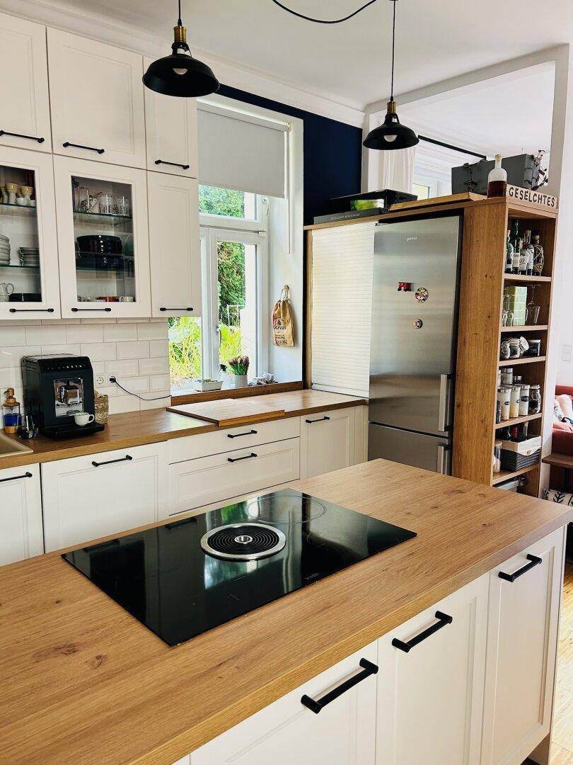 Gut ausgestattete Küche mit weißen Schränken, Holztheken, Kühlschrank und Blick ins Grüne durch das Fenster.