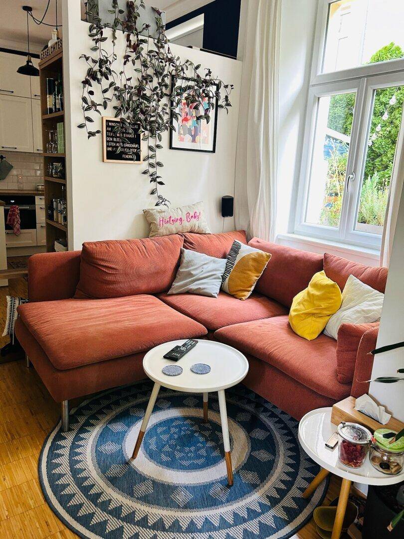 Gemütliches Wohnzimmer mit rotem Ecksofa, rundem Teppich und Blick in die offene Küche.