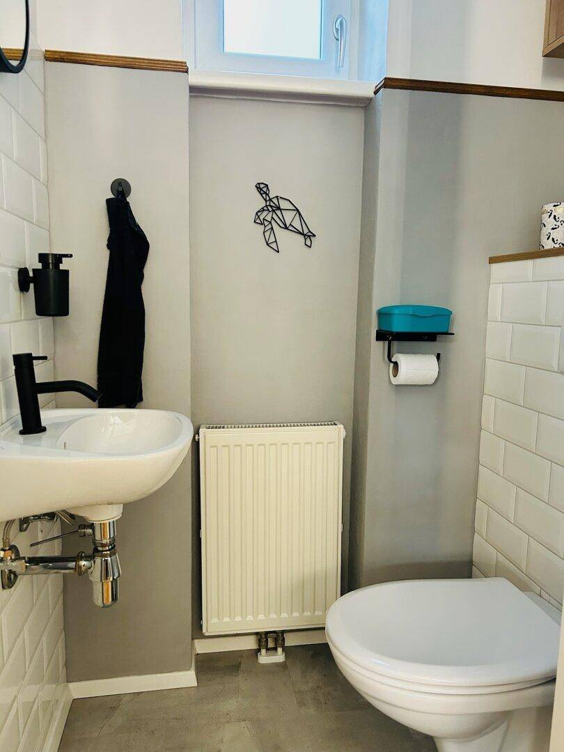 Modernes Gäste-WC mit weißer U-Bahn-Fliesenwand, kleinem Waschbecken und Heizkörper unter dem Fenster.