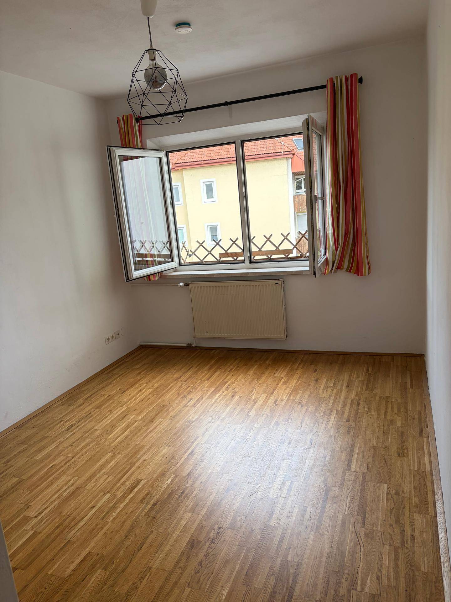 Leerer Raum mit Holzfußboden und einem geöffneten Fenster, das den Blick nach draußen freigibt.