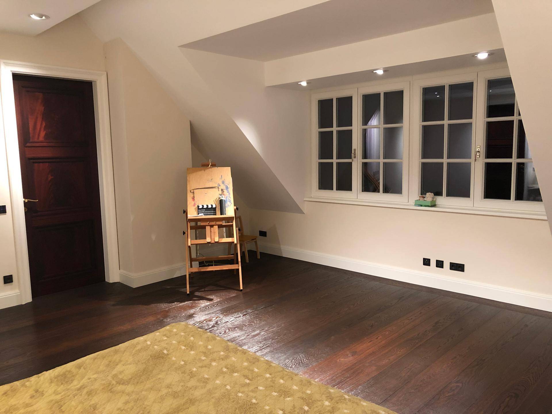 Helles Dachzimmer mit dunklem Holzboden, schrägen Wänden und großen Fenstern.