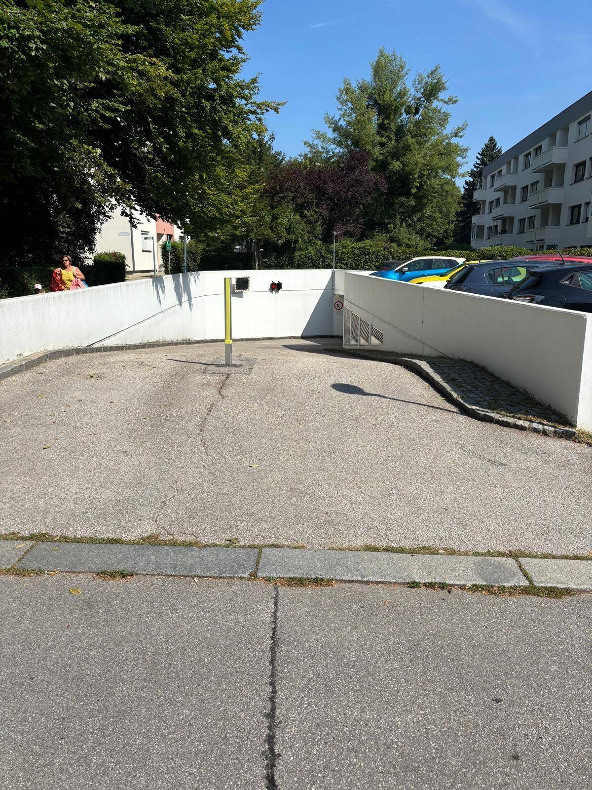 Außenansicht der Tiefgaragenzufahrt mit Rampe, umgeben von Bäumen und einem Wohngebäude.
