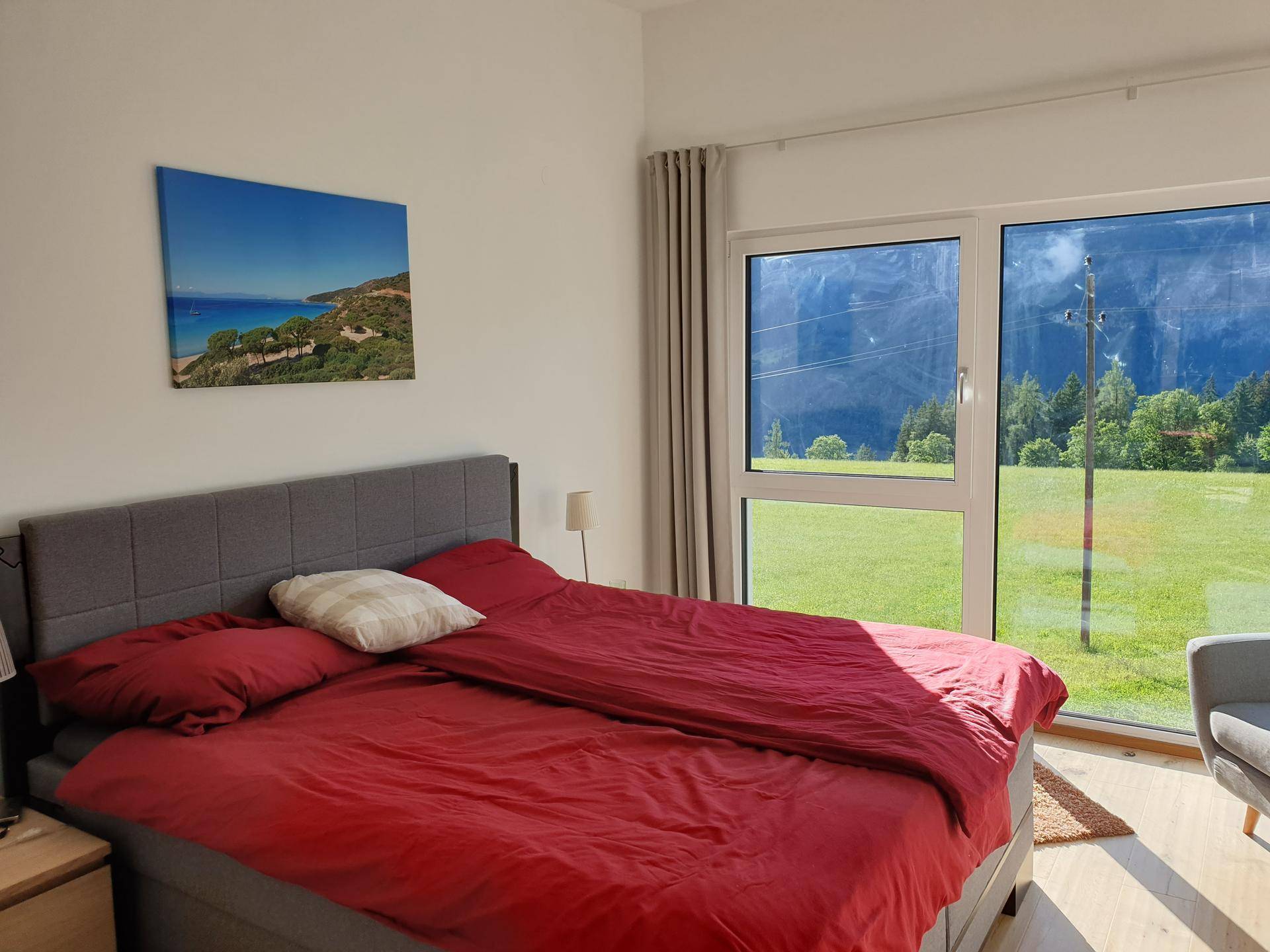 Helles Schlafzimmer mit großem Bett, roten Textilien und bodentiefen Fenstern mit Bergblick.
